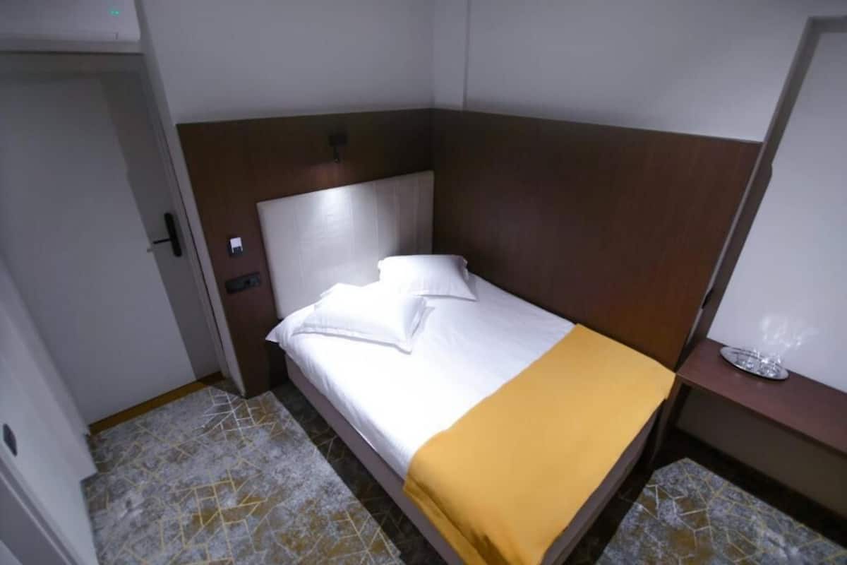 Une petite chambre d’hôtel avec un lit, une table de chevet et une lampe fixée au mur.