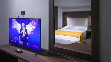 Habitación de hotel con una cama, un televisor mostrando 'Netflix Lucifer' y un control remoto sobre una mesa de madera.