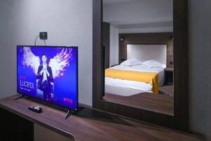 A hotel room with a bed, a TV displaying 'Netflix Lucifer', and a remote control on a wooden table.