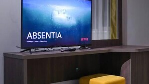 A flat-screen TV on a wooden console with a yellow ottoman and a mirror above it.