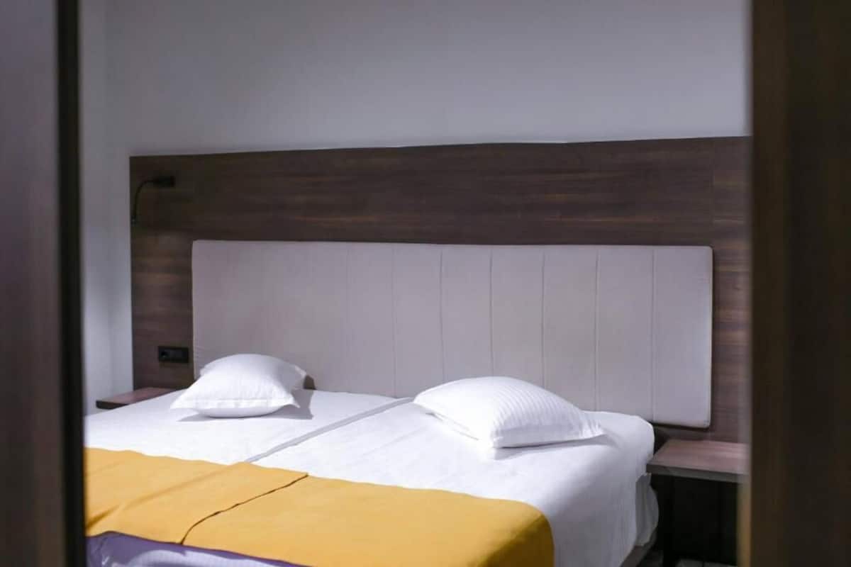 Une chambre d’hôtel avec une tête de lit en bois, deux lits avec du linge de lit blanc et un plaid jaune.