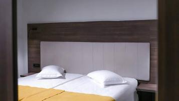 Habitación de hotel con cabecera de madera, dos camas con ropa blanca y una manta amarilla.