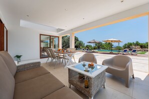 Ein moderner Außenbereich mit Tischplatte aus Glas, Korbstühlen und Blick auf einen Swimmingpool und Grünflächen.