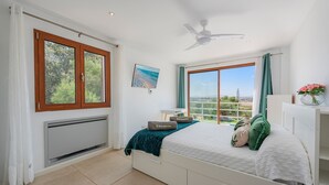 Een slaapkamer met een bed, een plafondventilator, een raam met gordijnen, een radiator en uitzicht op de omgeving.