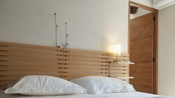 Ein Bett mit weißen Kissen und einer grauen Decke, ein hölzernes Kopfende, eine Nachttischlampe und ein Holzschrank.