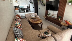 Ein modernes Wohnzimmer mit Sofa, Couchtisch und Fernseher. Im Hintergrund ist eine Küche erkennbar.