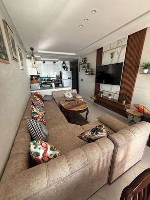 Ein modernes Wohnzimmer mit Sofa, Couchtisch und Fernseher. Im Hintergrund ist eine Küche erkennbar.