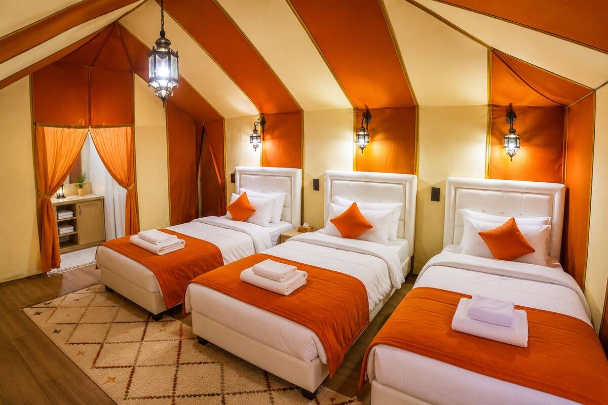 Ett hotellrum med tre sängar, alla med vita sängkläder och orange detaljer, ett litet skrivbord och en mönstrad matta.