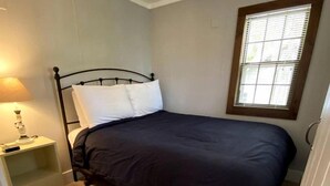 Une petite chambre avec un lit simple, un plancher en bois, une table de chevet avec une lampe et une fenêtre avec des stores.