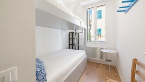 Een kleine kamer met een stapelbed, een klein rond tafeltje, een stoel, een radiator en een raam met luiken.