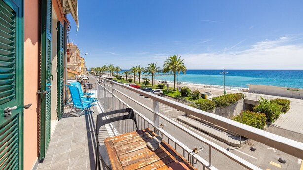 Een balkon met een houten tafel en stoelen, uitkijkend op een kuststraat met palmbomen en een strand.