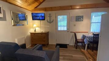 Una habitación acogedora con techo de madera, televisor montado en la pared, cómoda de madera, mesa de comedor con sillas y una puerta con ventanal.