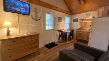 A cozy room with a wooden ceiling, a flat-screen TV, a wooden dresser, a gray sofa, and a kitchen area with wooden cabinets and a refrigerator.