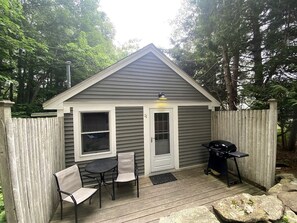 Une petite terrasse en bois avec une table et des chaises, un barbecue et un abri de jardin doté d’une porte et de fenêtres.