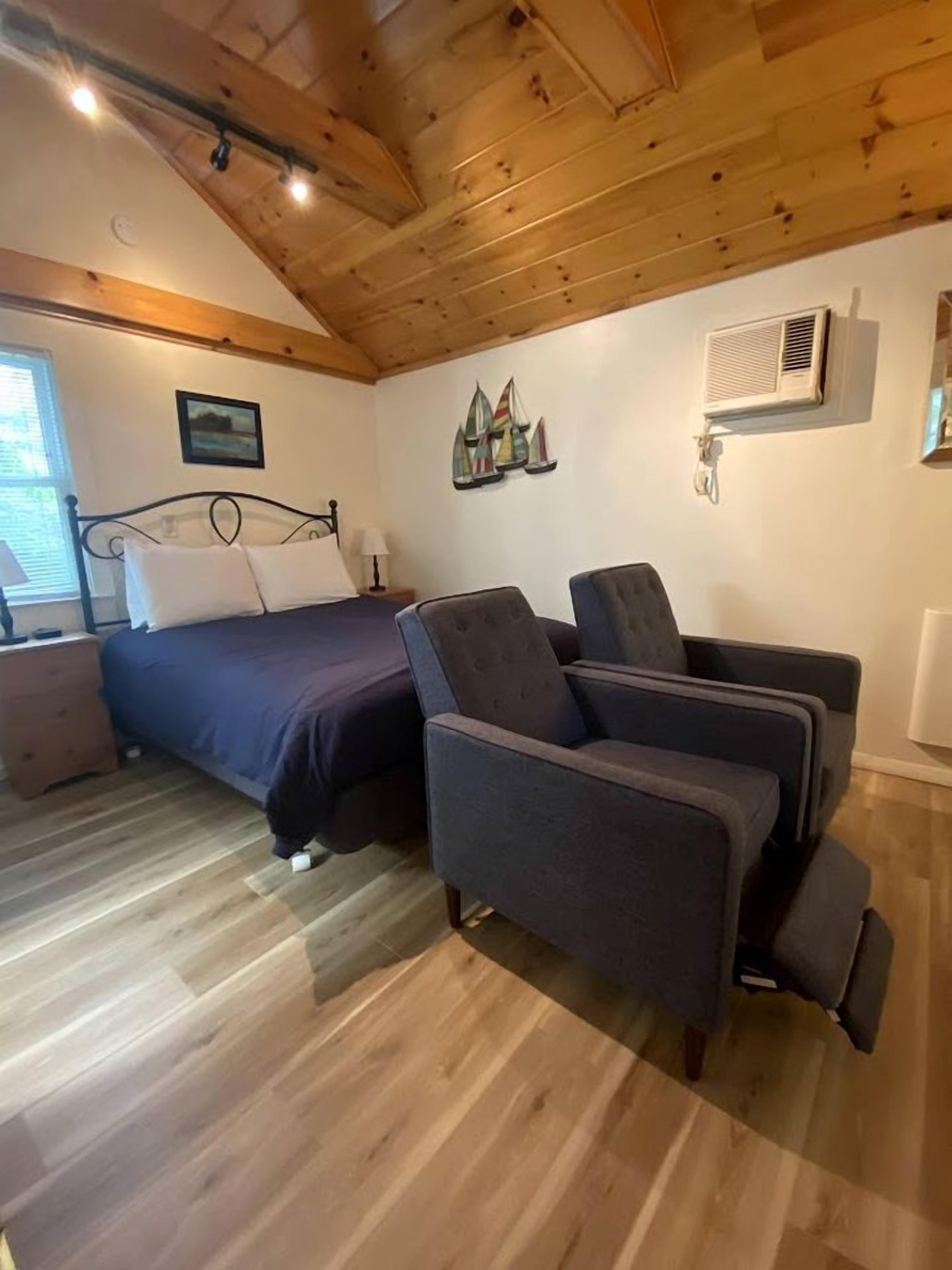 Une chambre à coucher comprenant un lit, deux fauteuils, un plafond en bois, un climatiseur fixé au mur et une fenêtre avec des stores.