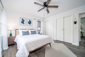Ein Schlafzimmer mit Bett, Deckenventilator, Nachttisch, Lampe, einem Fenster mit Jalousien und einer Tür.