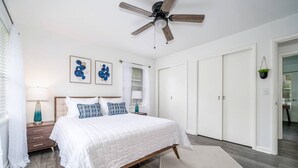 Une chambre à coucher comprenant un lit, un ventilateur de plafond, une table de chevet, une lampe, une fenêtre avec des stores et une porte.