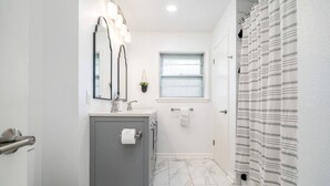 Une salle de bain avec un meuble gris, un miroir, un lavabo, une fenêtre et un rideau de douche.