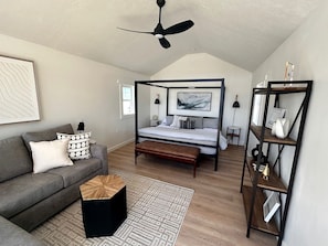 Ein Schlafzimmer mit einem Himmelbett, einem grauen Sofa, einem Holztisch, einem schwarzen Hocker, einem gemusterten Teppich und einem schwarzen Metallregal.