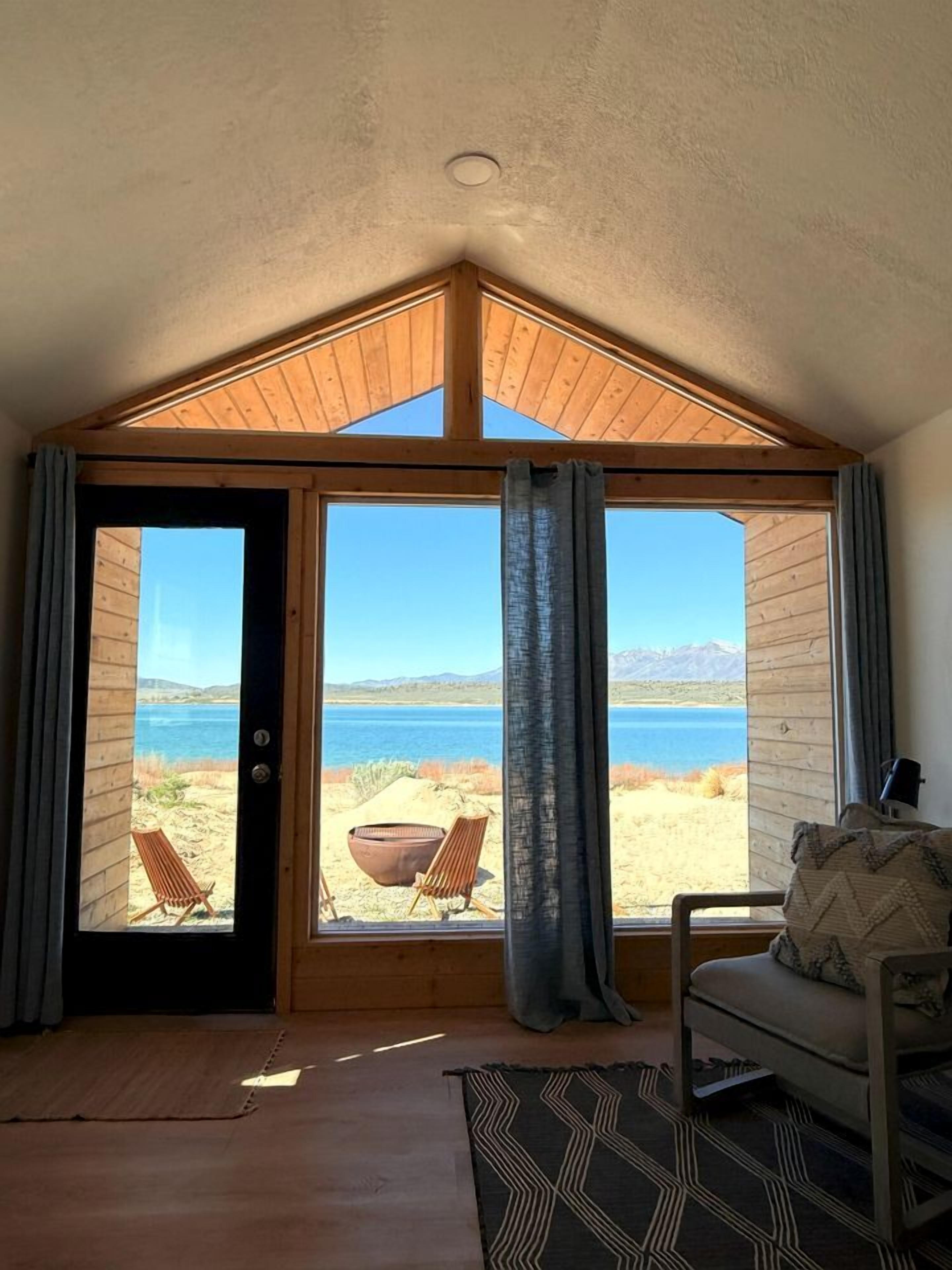 A room with a large window overlooking a beach and mountains, a wooden ceiling, and a chair with a cushion.