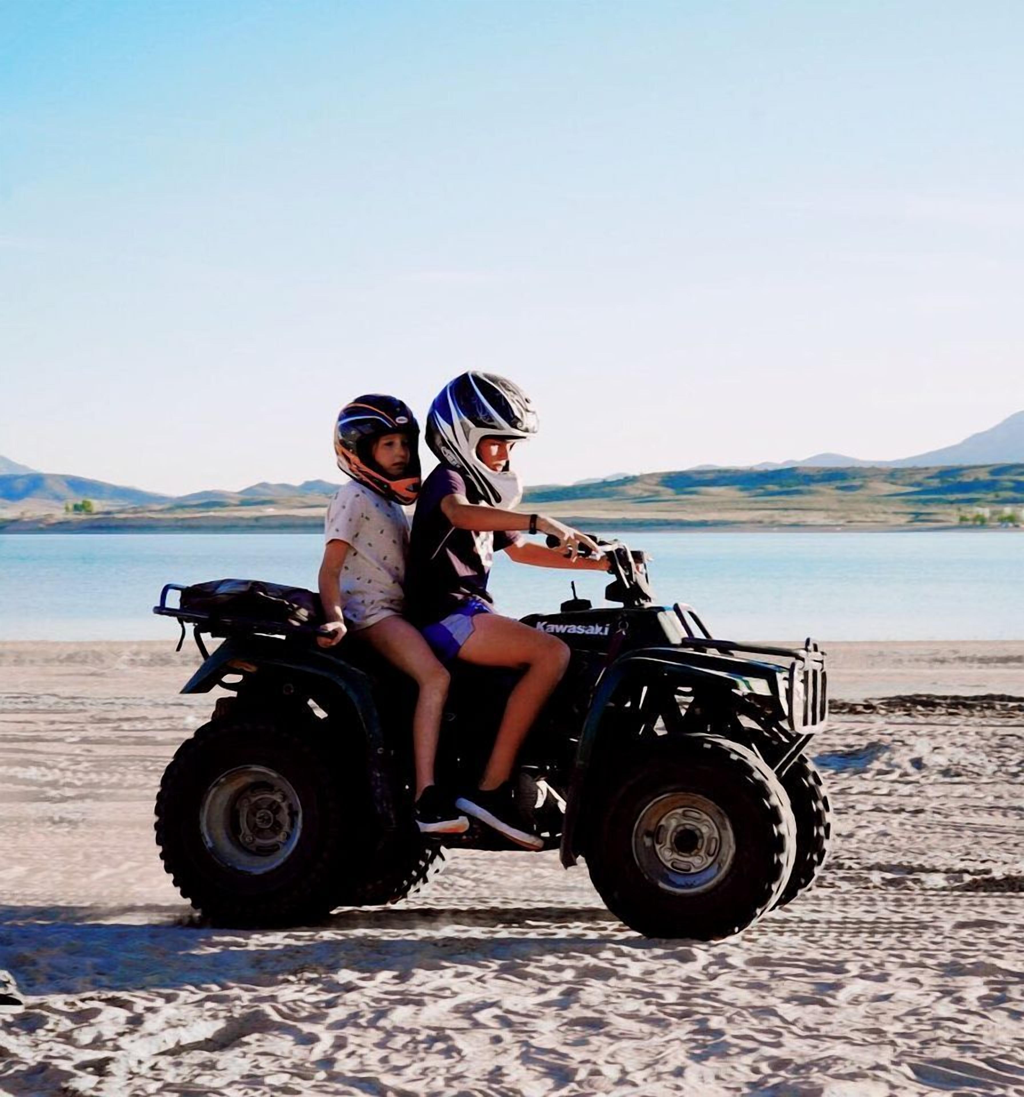 Two people on a four-wheeled vehicle on a sandy beach with a body of water and hills in the background.