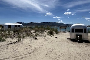 Dos remolques sobre una playa arenosa con un lago y montañas al fondo.