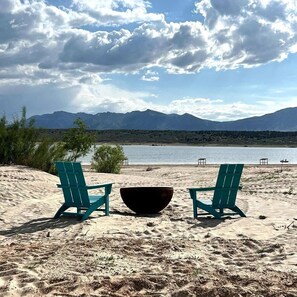 Une plage avec deux chaises vertes et un bol noir, un lac et des montagnes en arrière-plan.