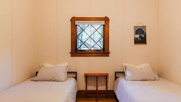 Ein Zimmer mit zwei Betten, einem kleinen Holztisch, einem Fenster mit Holzrahmen und einem an der Wand hängenden Bild.