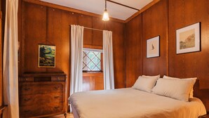 Una habitación con paneles de madera, una cama, un aparador, una ventana con cortinas e imágenes enmarcadas en las paredes.
