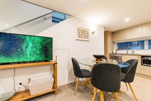 A modern kitchen with a dining area, a flat-screen TV, and a window with blinds. - Serene CBD Stay | 2BR | Balcony + Laundry (Christchurch)