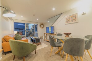 Ein modernes Wohnzimmer mit einer Couch, Sesseln, einem Glastisch, einem Fernseher und Blick durch eine Glasdoor.