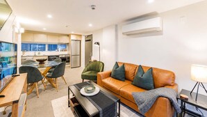 Ein modernes Wohnzimmer mit einer Couch, einem Esstisch und Stühlen. Im Hintergrund ist eine Küche mit Schränken und einem Kühlschrank zu sehen.