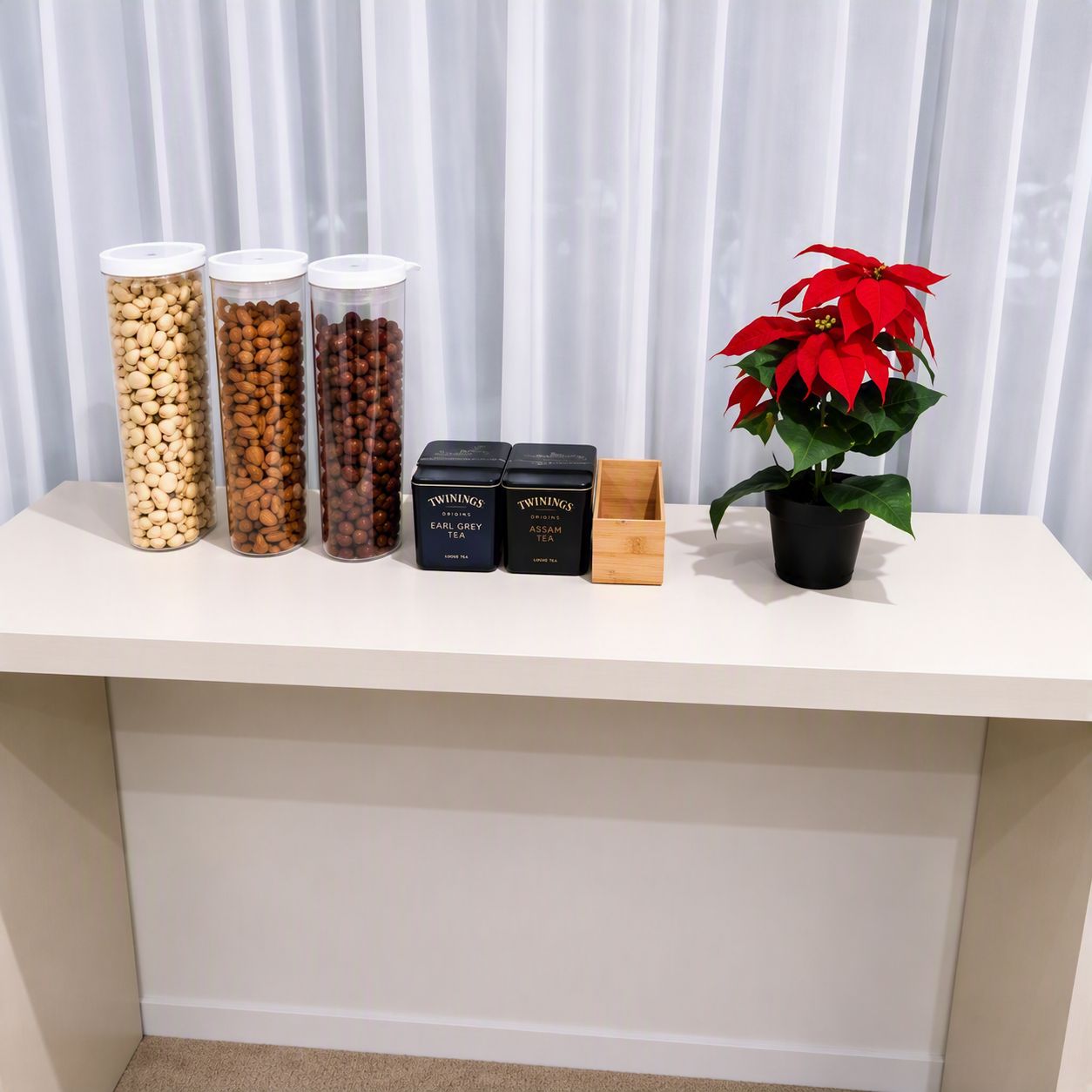 A white table with containers of nuts, two boxes labeled 'Twinings Earl Grey Tea' and 'Twinings Assam Tea,' and a potted red poinsettia plant.