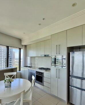 A modern kitchen with stainless steel appliances, a dining table with chairs, and a view of the city through large windows. - Sydney olympic park Free parking 08 (Sydney Olympic Park)