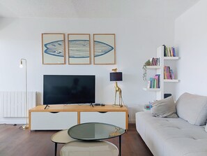A living room with a TV, a glass coffee table, a sofa, a desk with a lamp, and bookshelves. - Apartment Casino by Interhome (Lacanau)
