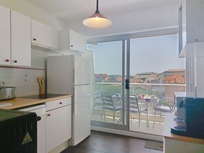 Une cuisine avec des meubles blancs, un réfrigérateur et une vue sur la ville, visible à travers une porte-fenêtre coulissante.