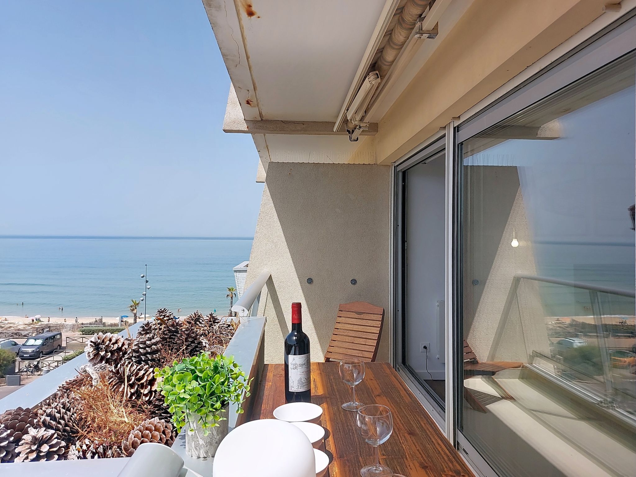 Een balkon met een tafel, een fles wijn en glazen, met uitzicht op een strand en de oceaan.