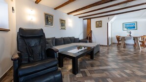 Uma sala de estar com poltrona de couro, sofá e mesa de centro com um jogo americano. Escada de madeira leva a outro cômodo, e há uma área de jantar com mesa e cadeiras.