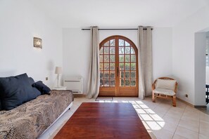 A bedroom with a large window, a bed with patterned bedding, a bedside table with a lamp, and a chair.