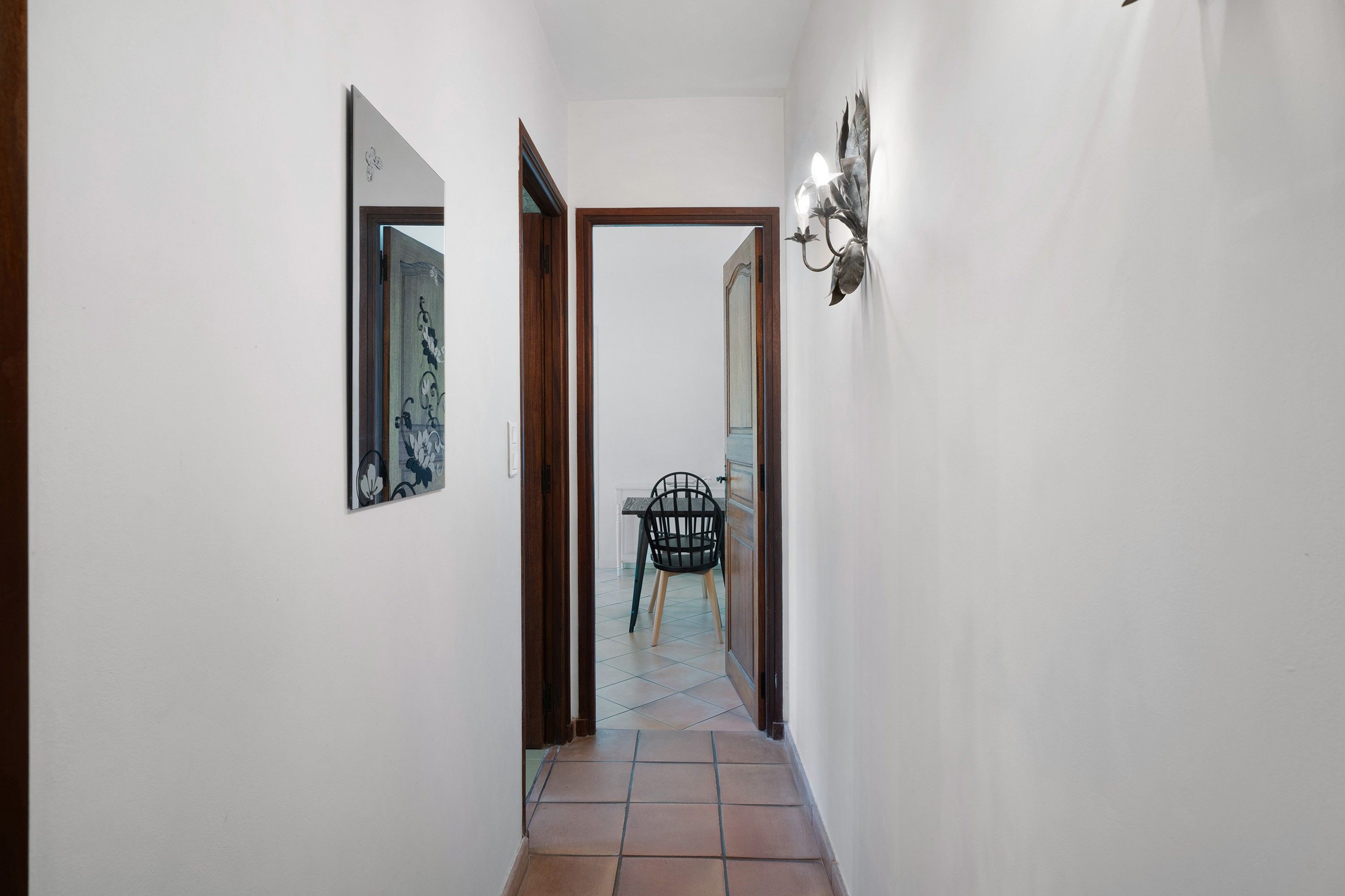 A narrow hallway with a tiled floor, a door leading to a room with a chair and a table, and a wall-mounted light fixture.