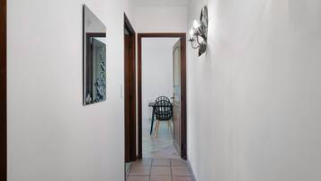 A narrow hallway with a tiled floor, a door leading to a room with a chair and a table, and a wall-mounted light fixture.