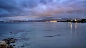 Paesaggio costiero con acque calme, una linea di costa lontana con edifici e una catena montuosa sullo sfondo.