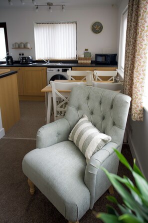 Un salon avec un fauteuil gris, un coussin à rayures, une table à manger et, au fond, une cuisine.
