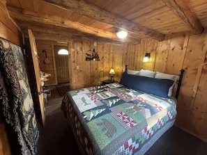 A wooden cabin bedroom with a bed, a nightstand, a lamp, a door, and a wall clock.