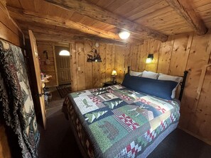 A wooden cabin bedroom with a bed, a nightstand, a lamp, a door, and a wall clock.