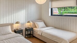 Ett sovrum med två sängar, ett fönster med utsikt över grönområden och ett nattduksbord med en lampa.