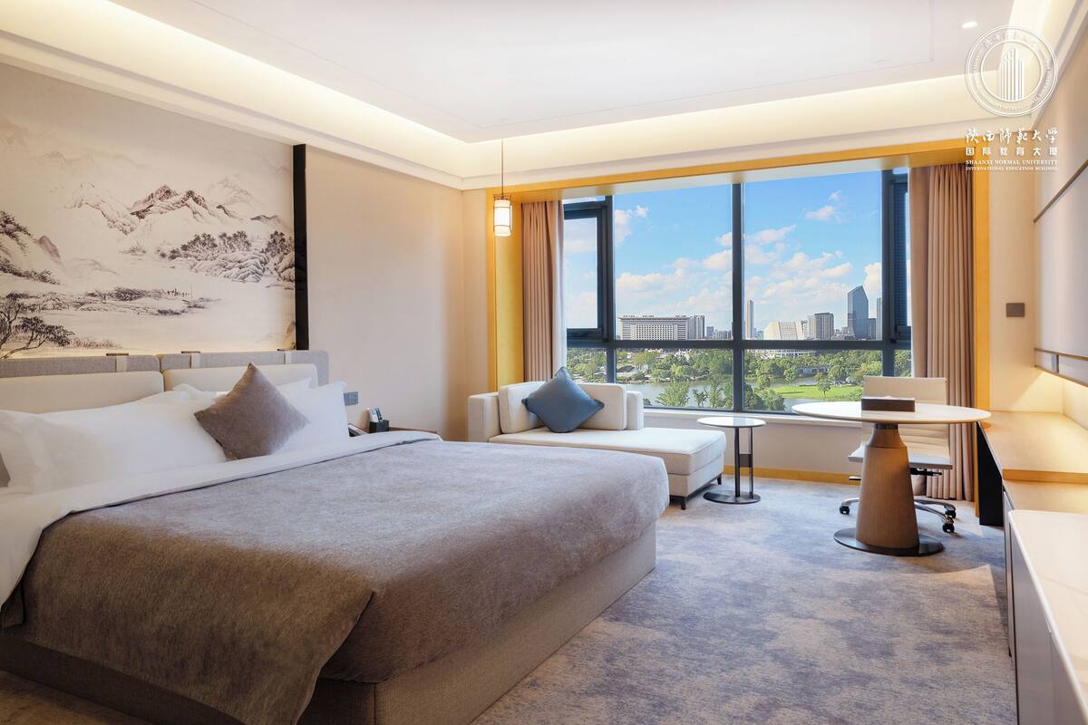 A hotel room with a large bed, a sofa, a small table, and a view of the cityscape.