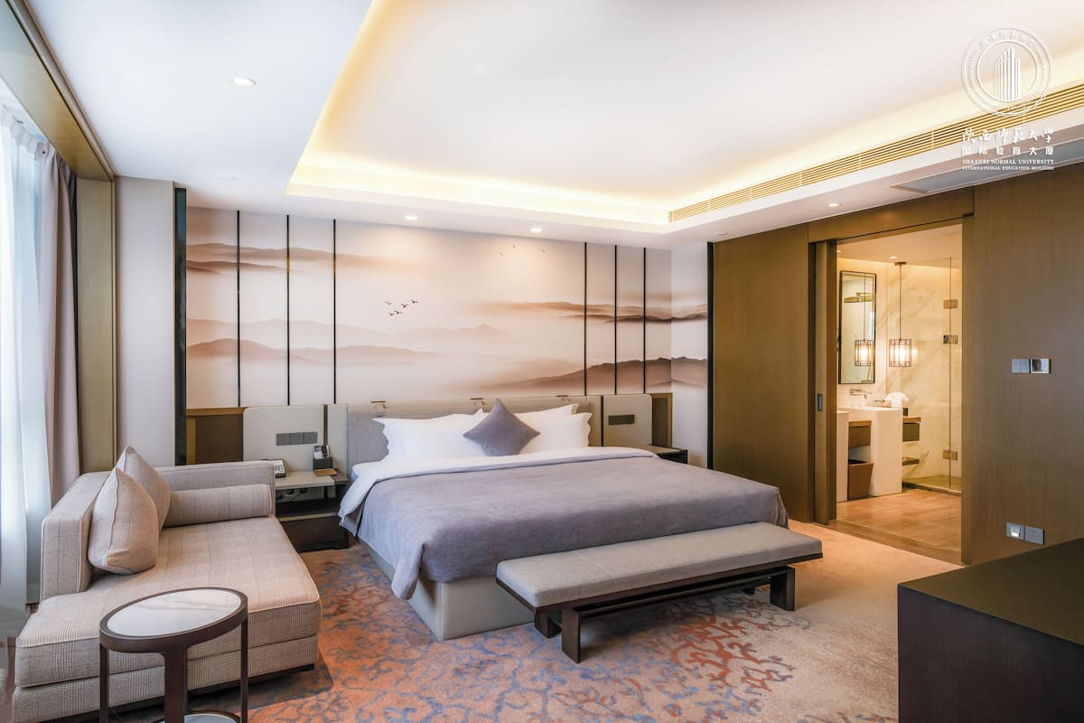 A modern hotel room with a large bed, a sofa, a bench, and a desk.