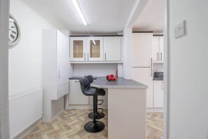 Eine kleine Küche mit weißen Schränken, einem schwarzen Barhocker und einer Obstschale auf der Arbeitsfläche.