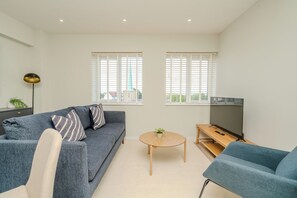 A modern living room with a sofa, a round coffee table, a TV, and two windows with blinds. - Church House by Guestz (Filton)